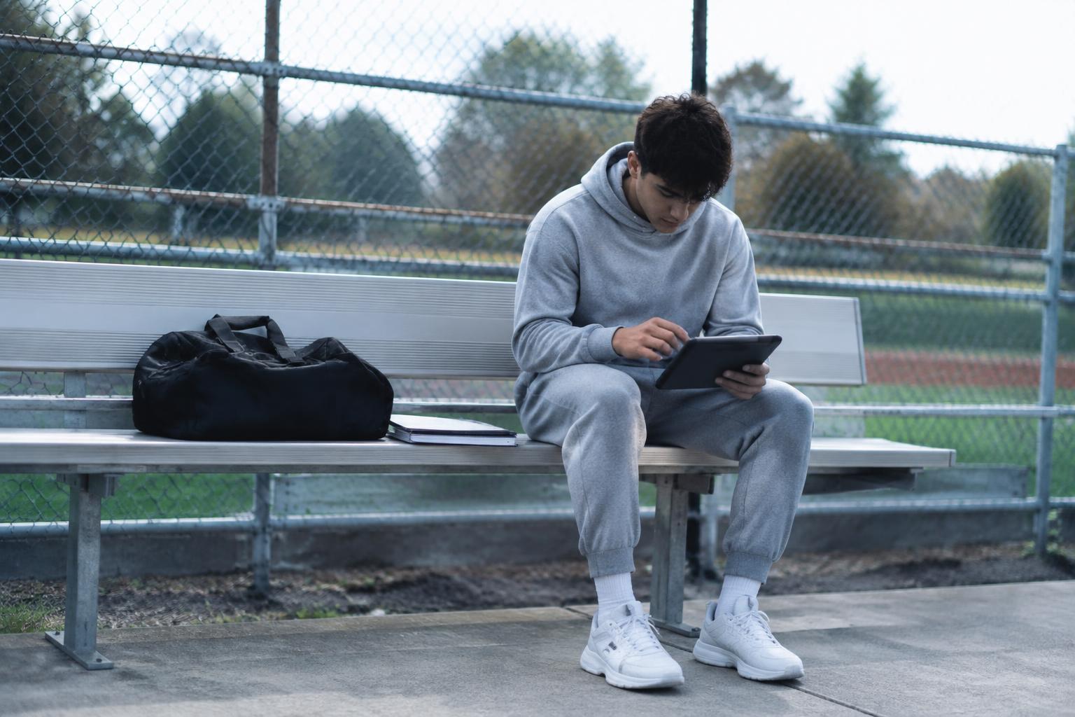 Giovane atleta in tuta sportiva che studia sul tablet seduto a bordo campo con la borsa sportiva accanto